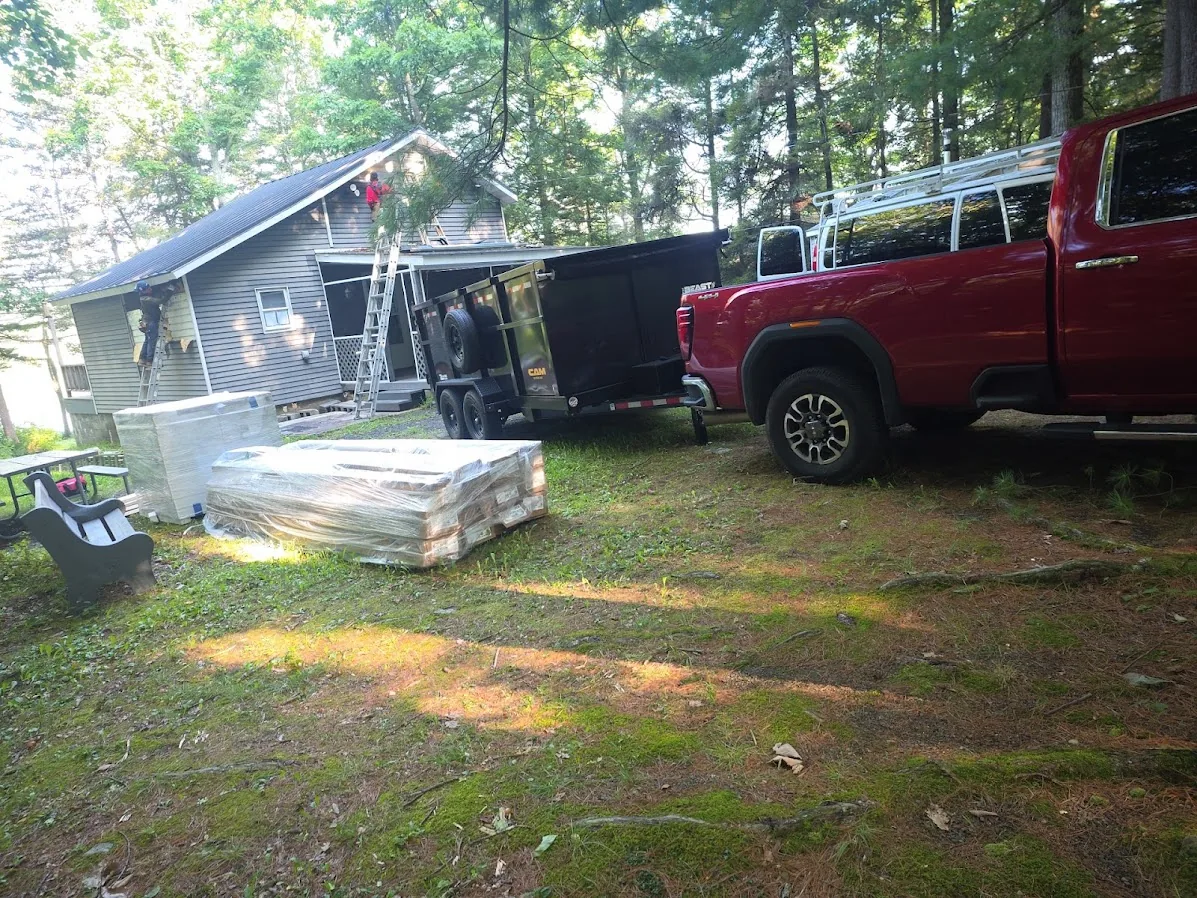 Dump trailer for the old roof and wrapped pallets of new roofing material staged beside a Poconos home