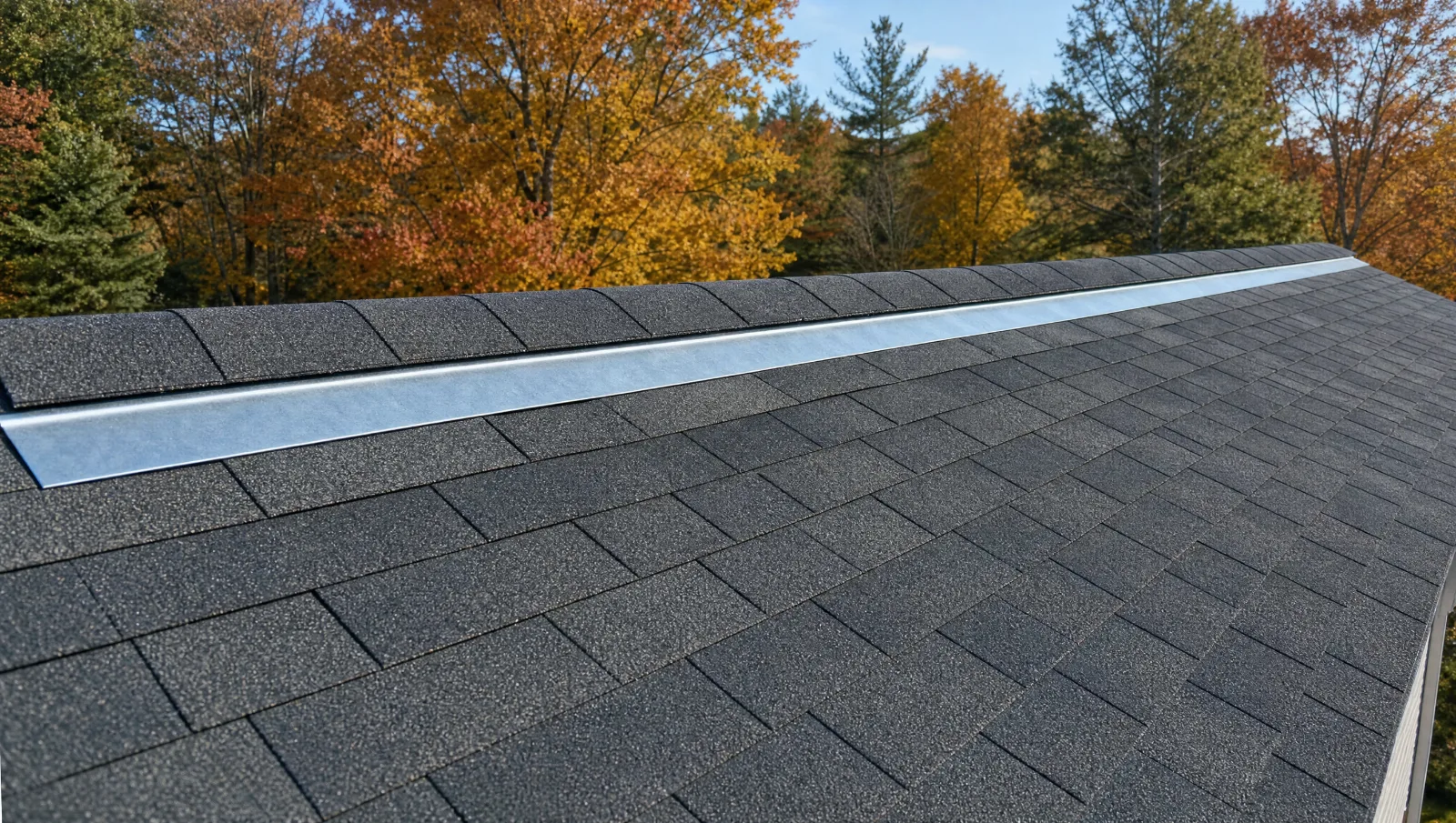 Zinc strip installed along the full length of a shingle roof just below the ridge cap, with fall trees behind it