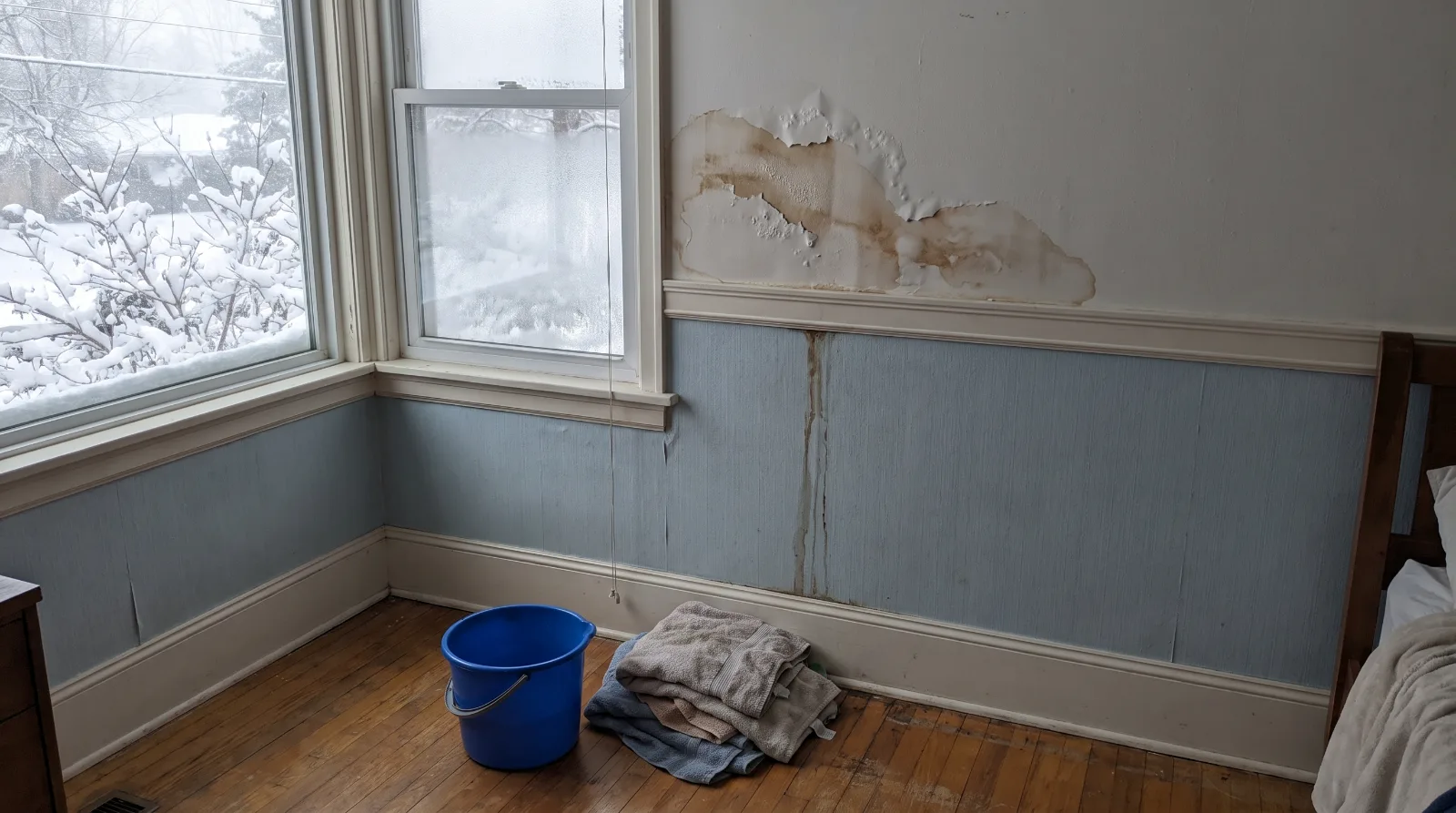 Water stain and peeling paint at the top of a bedroom window on an exterior wall, with a bucket and towels below